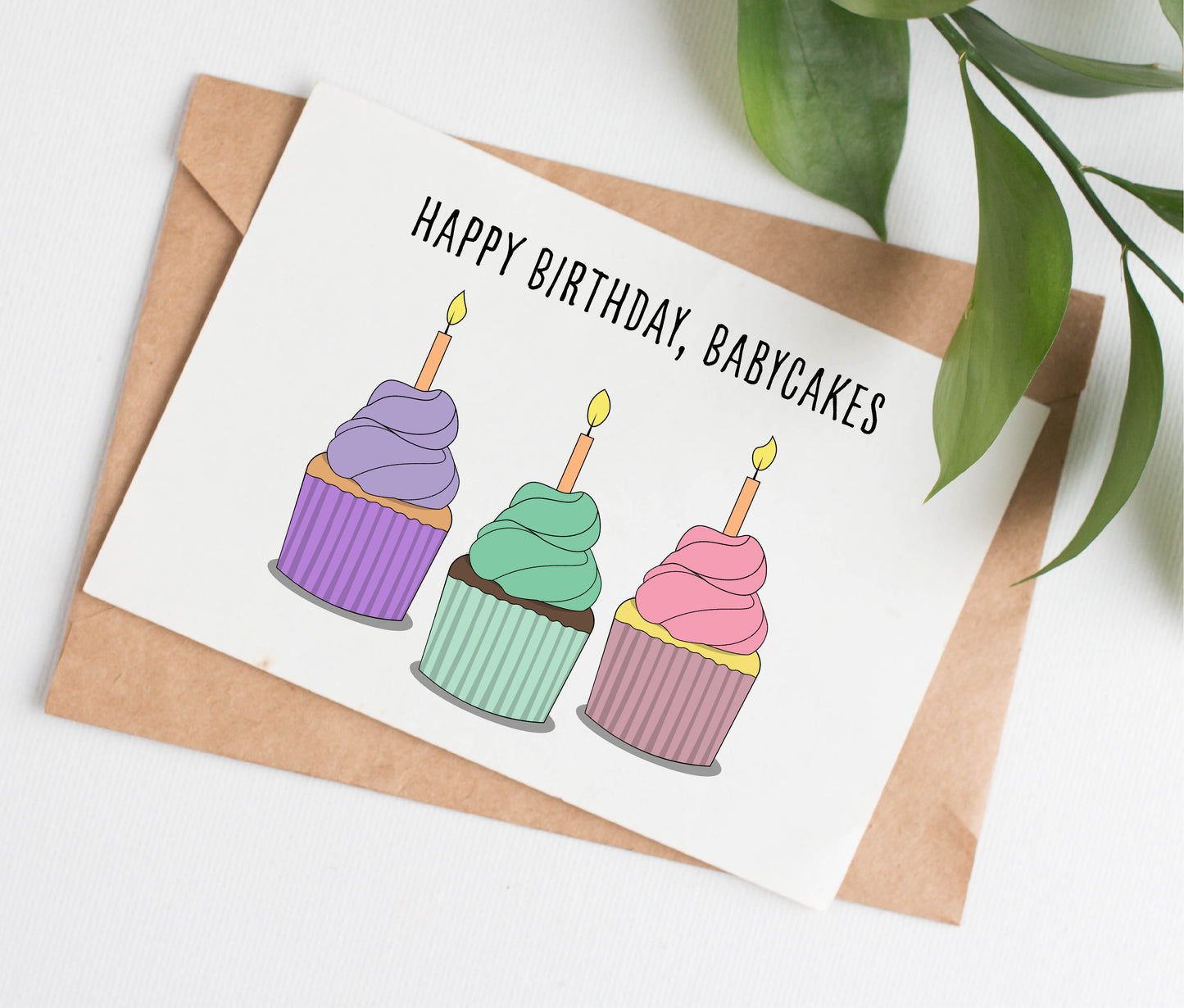 Handmade greeting card with horizontal design of three illustrated birthday cupcakes in pastel colors. Caption on card reads "HAPPY BIRTHDAY, BABYCAKES." Card is shown with a kraft brown paper envelope.