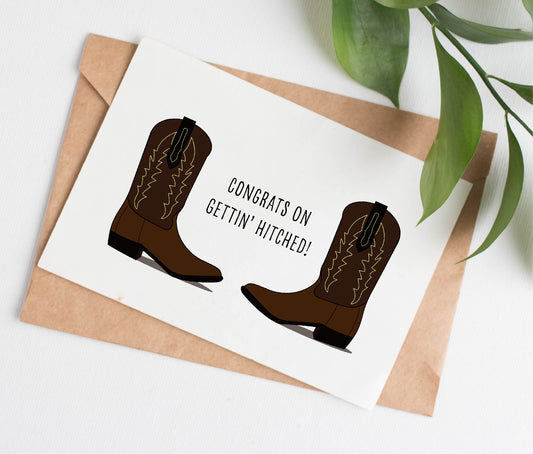 This wedding card is handmade in Canada. It shows two identical cowboy boots facing each other. The text between them says "Congrats on gettin' hitched!"